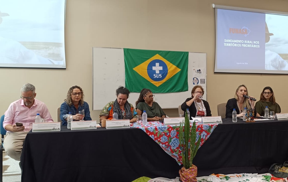 Pessoas sentadas à mesa em reunião do Grupo da Terra, com bandeira do SUS ao fundo e tela projetada.