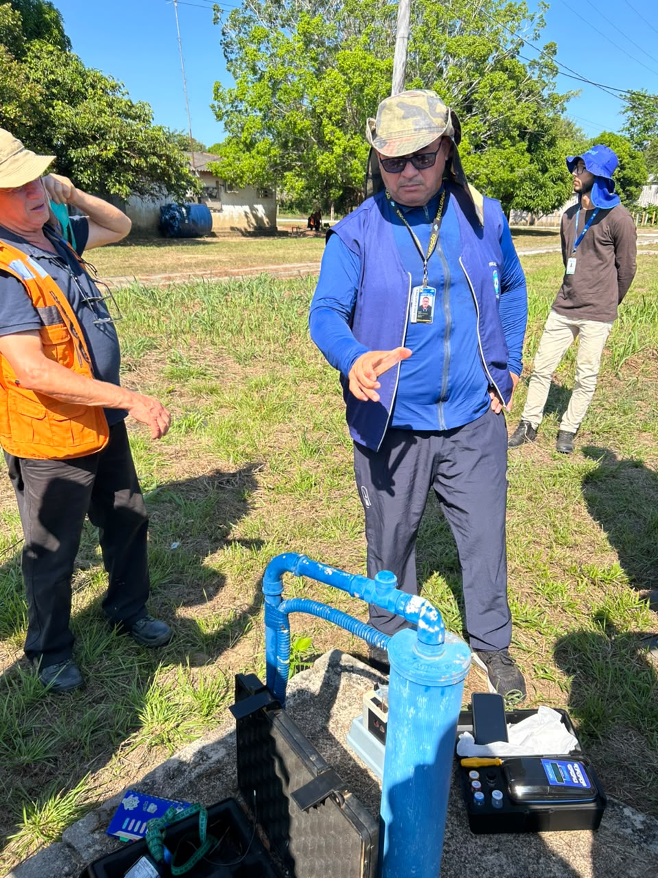 Três técnicos ao ar livre inspecionam tubulação de água azul e equipamentos em campo.
