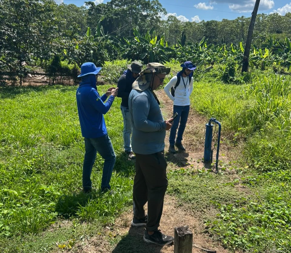 Técnicos de chapéu observam um poço de água em área rural gramada.