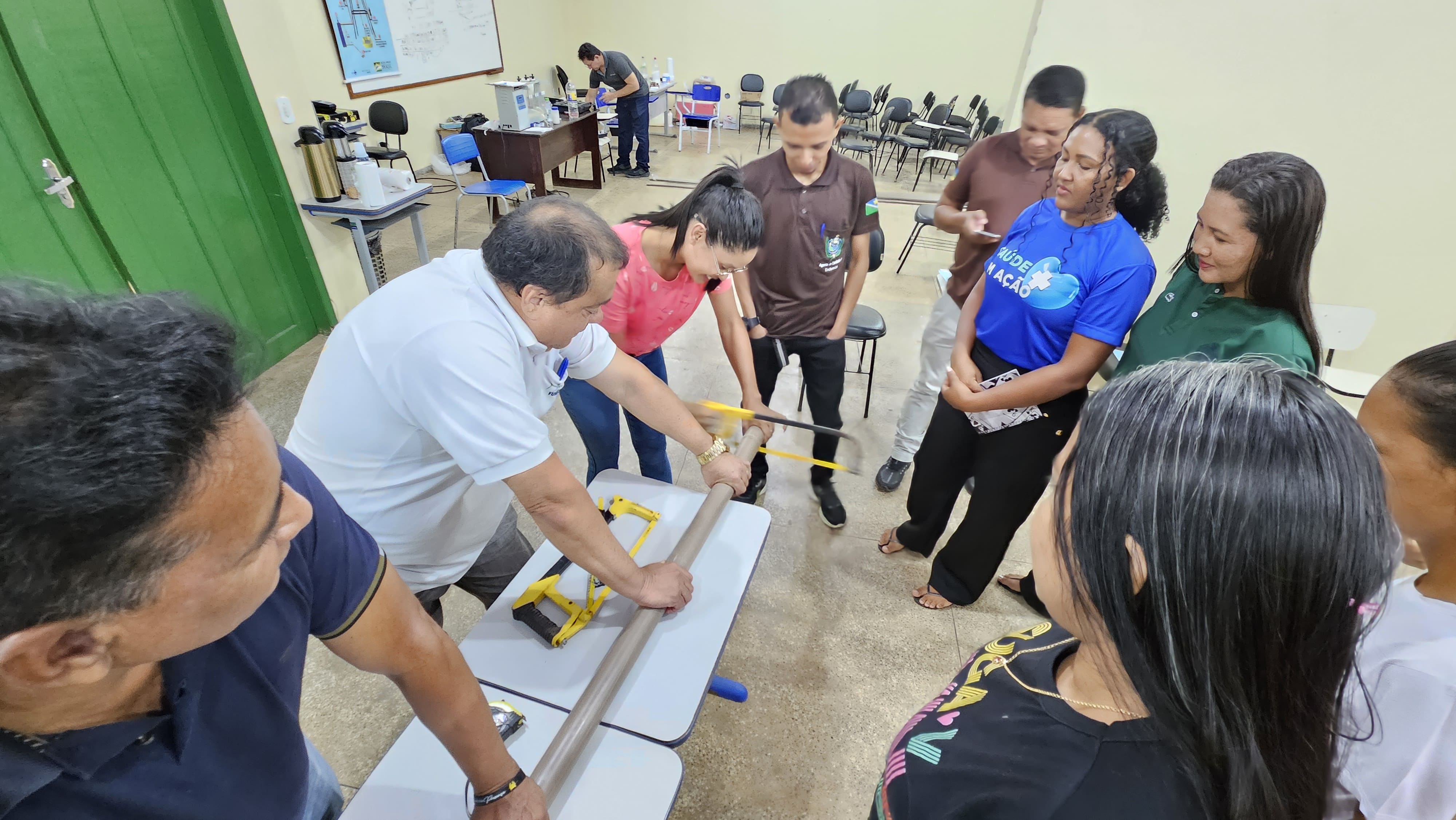 Instrutor orienta participantes que observam o corte manual de um cano de PVC com arco de serra na oficina.