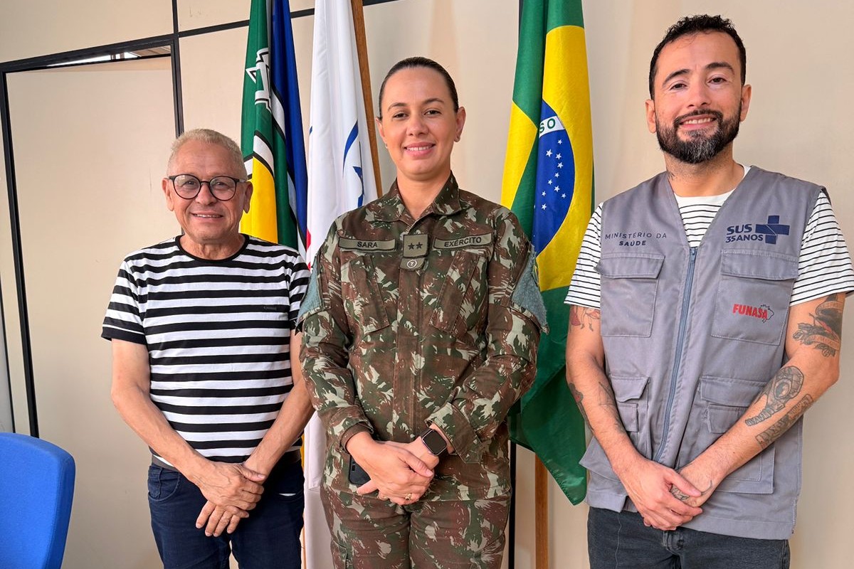 Uma militar do Exército e dois homens em pé lado a lado, em frente a bandeiras num escritório.