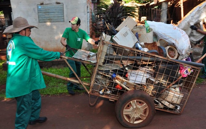 Trabalhadores de uniforme verde empurram carrinho metálico carregado de recicláveis.