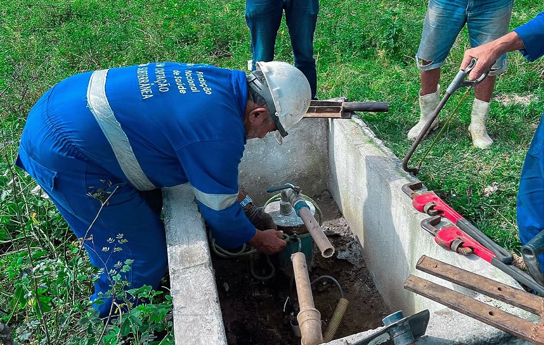Técnico de uniforme azul realiza manutenção em tubulações na caixa de concreto de um poço.