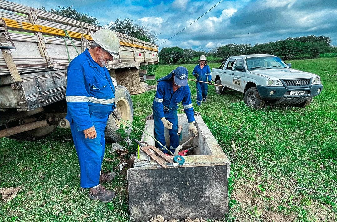 Trabalhadores fazem manutenção em caixa de poço ao lado de caminhão e picape em área rural.
