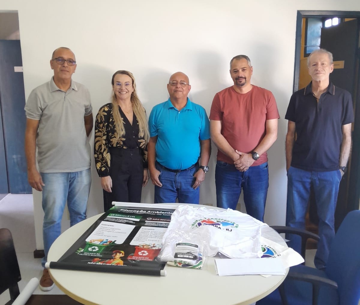 Cinco pessoas em pé atrás de mesa redonda com materiais informativos de educação ambiental em reunião.