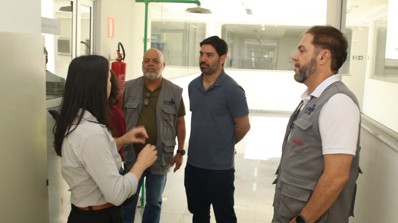 Mulher orienta três homens de colete e camisa polo no corredor de um laboratório.
