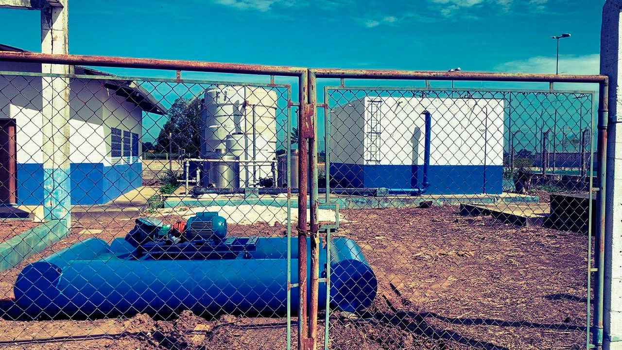 Instalação com bomba d'água azul e tanques ao fundo, vista através de uma cerca de metal.