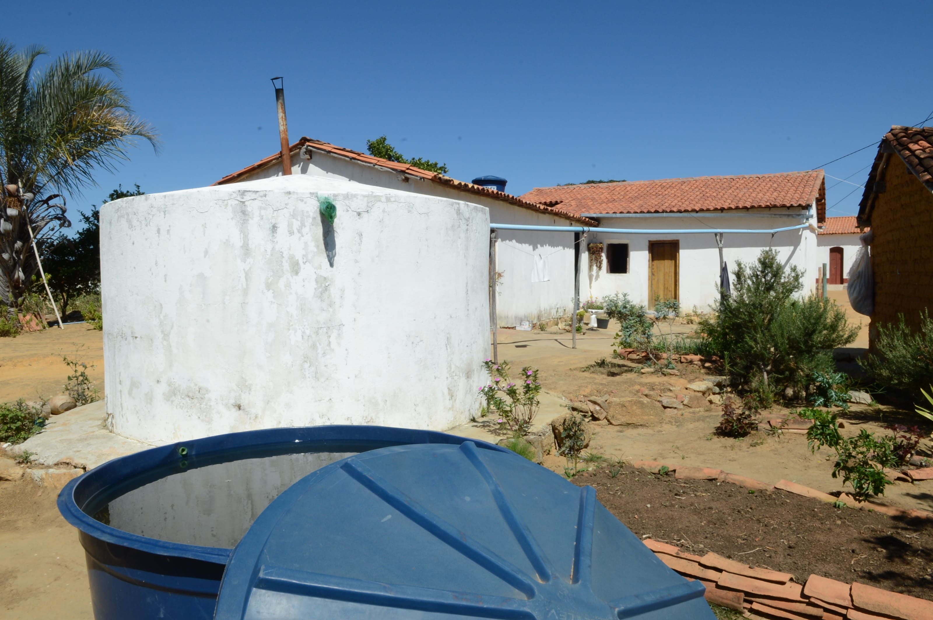 Cisterna branca de alvenaria e caixa d'água azul aberta em primeiro plano num quintal no semiárido.
