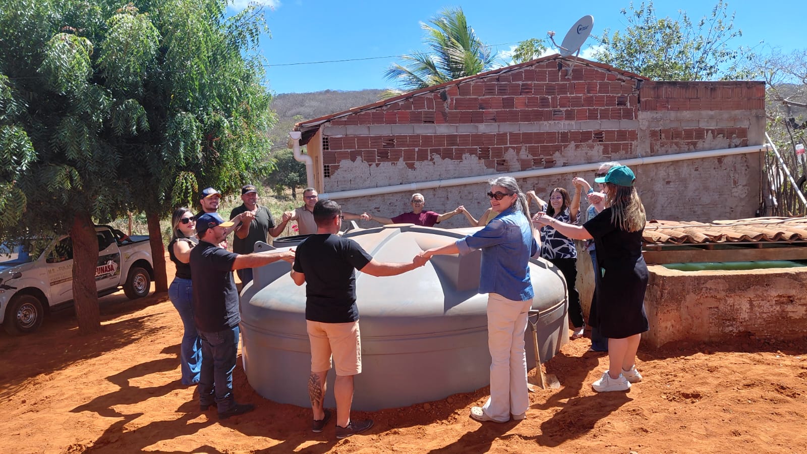 Pessoas de mãos dadas formando uma roda ao redor de uma cisterna cinza em local aberto.