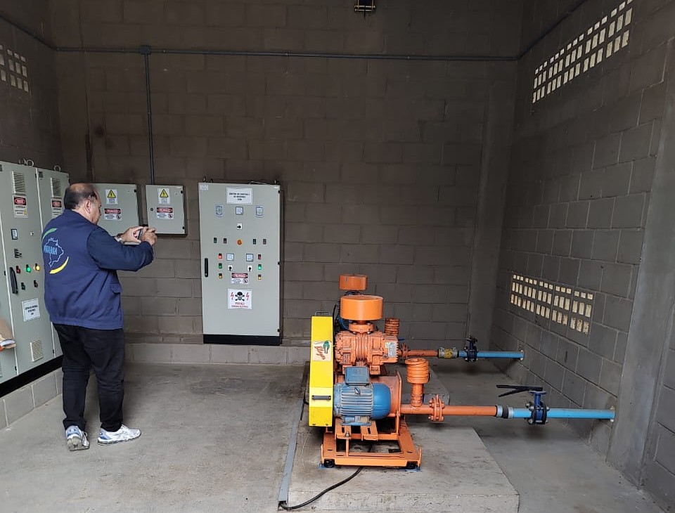 Homem de jaqueta azul fotografa sala de máquinas com painéis elétricos e motor com tubulações.
