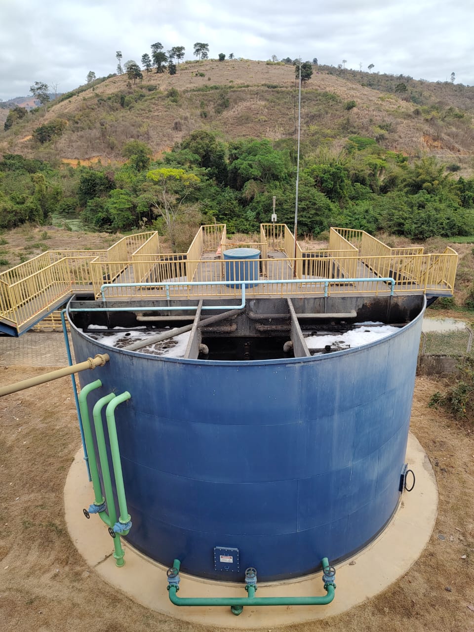 Tanque azul de tratamento de água ao ar livre com passarelas amarelas no topo e morro ao fundo.