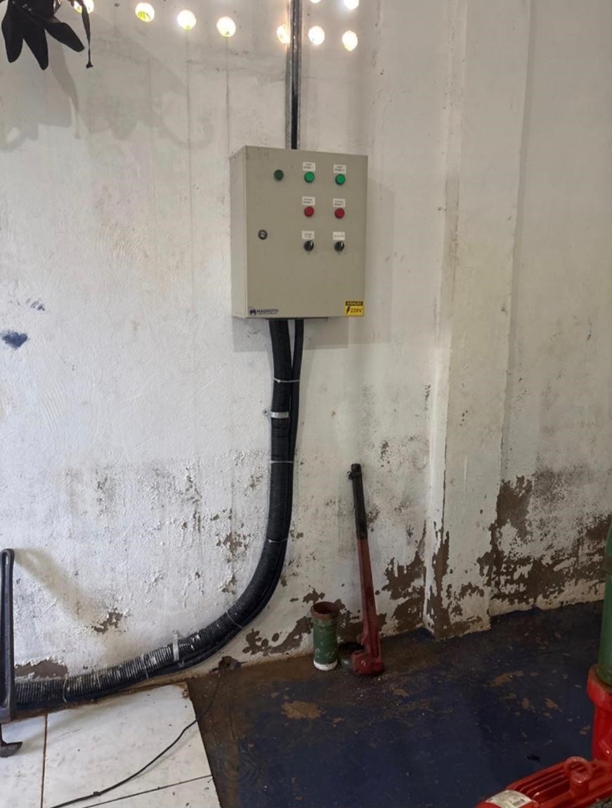 Painel elétrico cinza com botões e luzes coloridas fixado em parede rústica, com cabos e canos