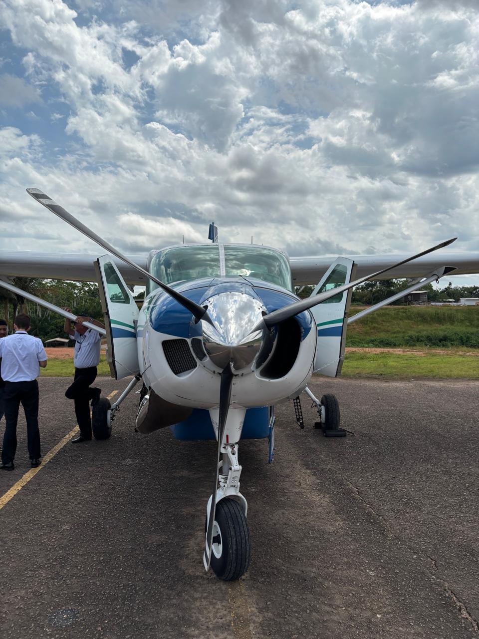 Avião monomotor, branco e azul, na pista com portas abertas e dois homens uniformizados ao lado.