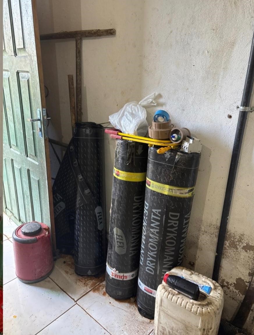 Rolos de manta asfáltica preta e outros materiais de construção armazenados em canto de parede.