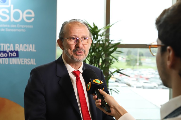 Homem de terno e gravata vermelha dá entrevista segurando microfone de TV, com banner institucional ao fundo.