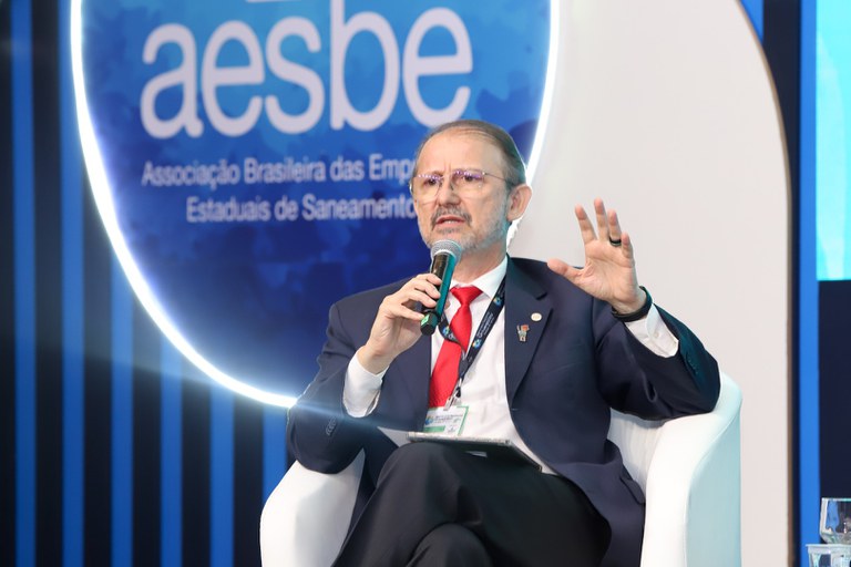 Homem de terno fala ao microfone sentado em poltrona branca durante painel do evento da Aesbe.