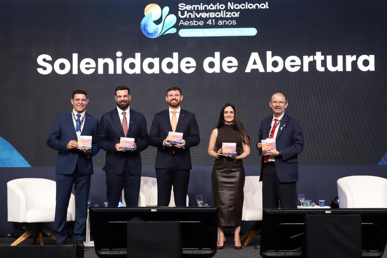 Cinco participantes posam no palco segurando publicações na solenidade de abertura do Seminário Universalizar 2025.