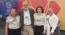 Quatro pessoas posam lado a lado em evento institucional, usando crachás; ao fundo, painéis coloridos e banner, uma delas segura bolsa da Funasa.