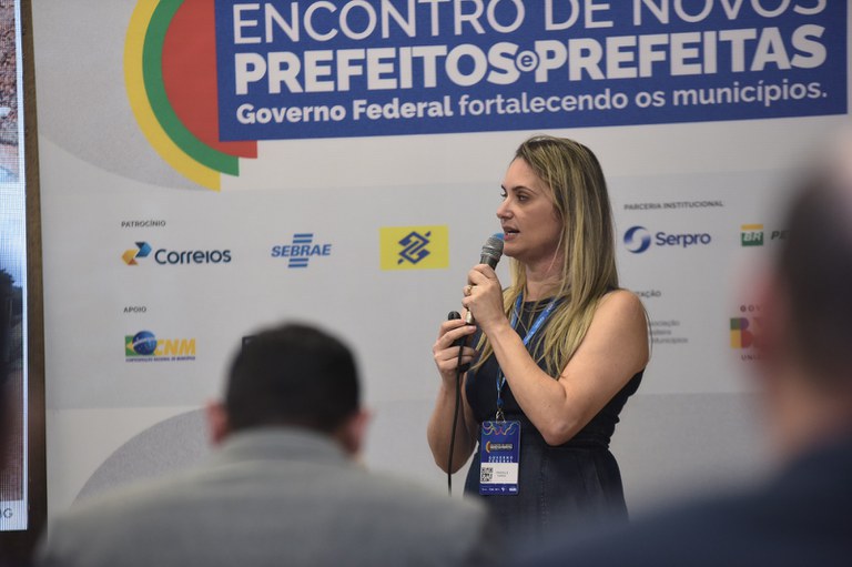 Mulher em pé, falando ao microfone em palco de evento institucional, com painel ao fundo escrito “Encontro de Novos Prefeitos e Prefeitas”.