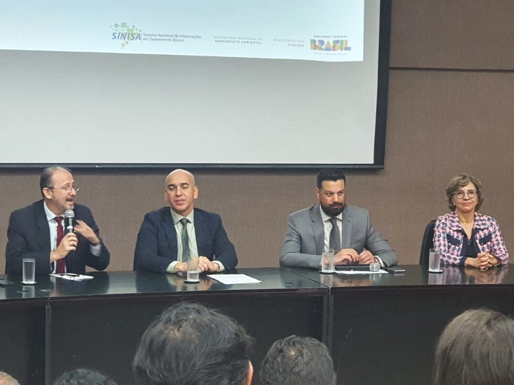 Mesa com quatro autoridades sentadas em auditório, durante evento institucional, com microfones, copos de água e público à frente.