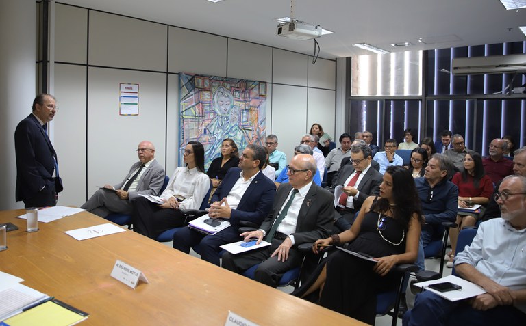 Reunião institucional com participantes sentados em auditório, ouvindo palestrante em pé. Ambiente formal, com mesa, cadeiras, quadro artístico na parede e projetor no teto.