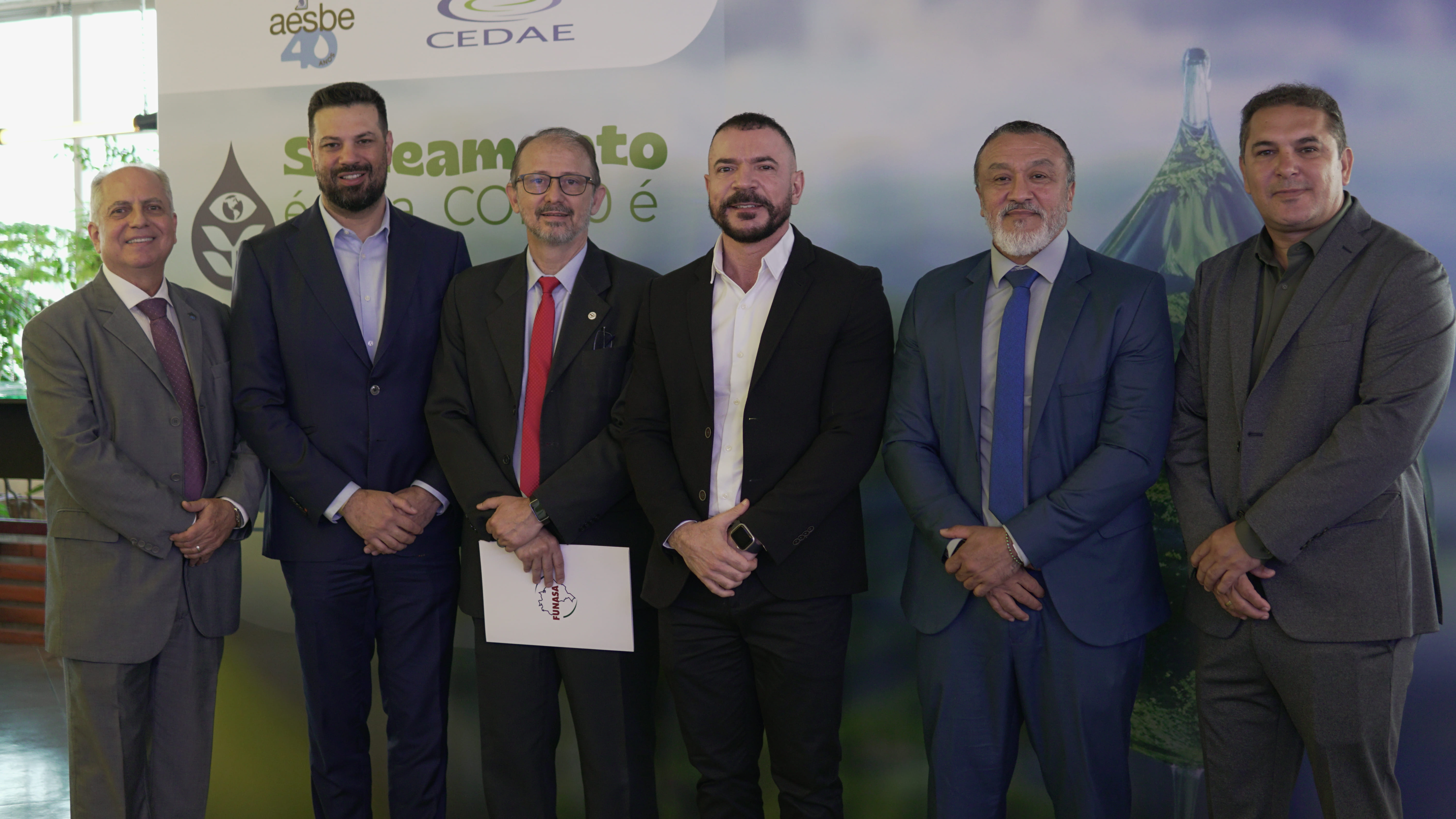 Seis homens em trajes formais posam lado a lado em evento institucional da Aesbe, diante de painel com logos e temática de saneamento.