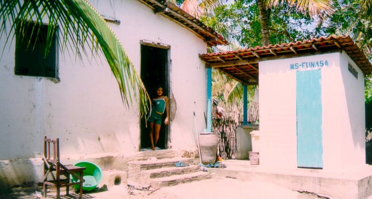 Mulher em pé na porta de uma casa simples em área rural, com coqueiros ao redor; ao lado, pequeno banheiro externo identificado como MS-FUNASA.