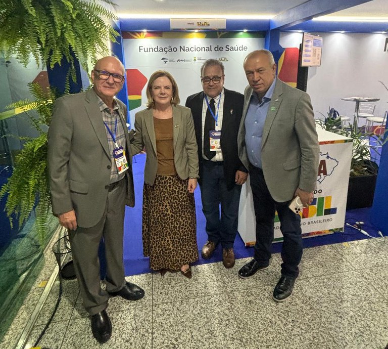 Quatro pessoas posam sorrindo em frente ao estande da Funasa, sobre piso de granito e cercadas por plantas.