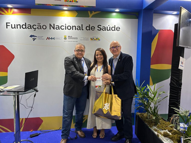 Duas autoridades entregam material institucional a mulher em estande da Funasa durante evento