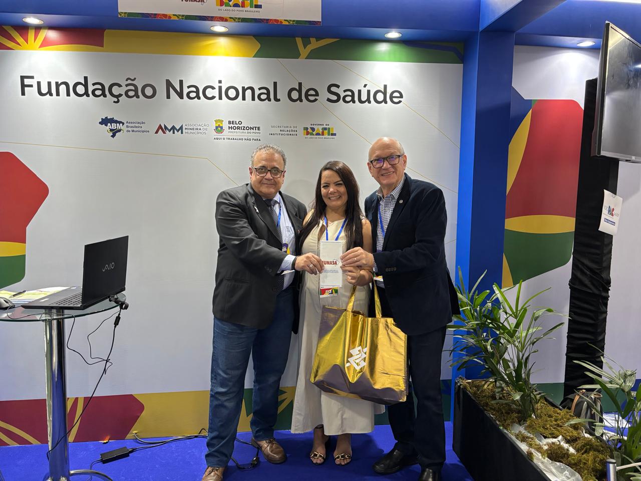 Duas autoridades entregam material institucional a mulher em estande da Funasa durante evento