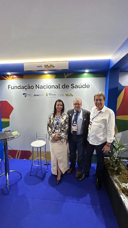 Três pessoas posam em estande da Funasa em evento nacional, com painel institucional e piso azul