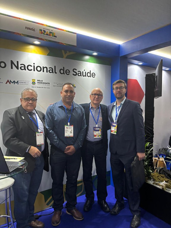 Quatro homens posam em estande institucional da Funasa durante evento, com credenciais e painel oficial ao fundo