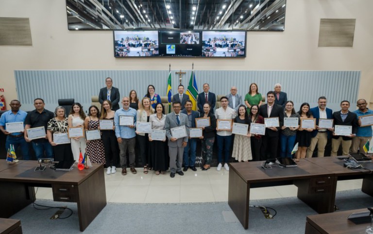 Grupo de pessoas em salão oficial segurando certificados, alinhadas em frente a mesas e bandeiras do Brasil e estados, sob painel de telas no alto.