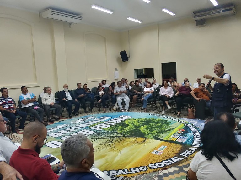 Palestrante fala com microfone para grupo em roda, em sala ampla, com grande banner colorido no centro do chão.