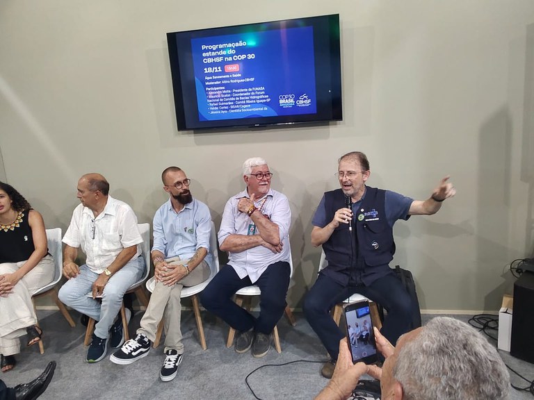 Palestrantes sentados em linha; um deles fala ao microfone enquanto outro grava com o celular.