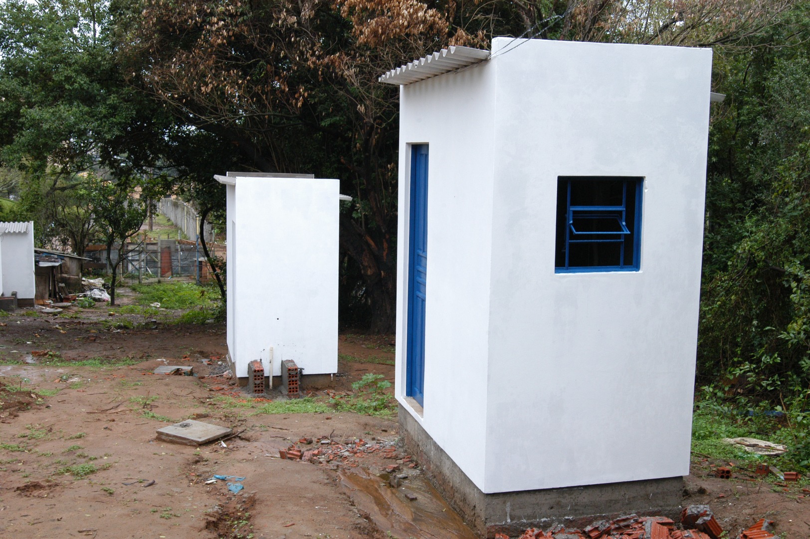 Banheiros brancos recém-construídos com portas azuis em área externa de solo úmido e árvores ao fundo.