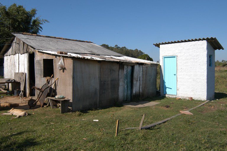 Casa rústica de madeira com banheiro branco e porta azul ao lado, em área rural ensolarada.