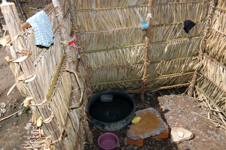 Estrutura simples de folhas trançadas formando um banheiro improvisado com bacia d’água no chão.