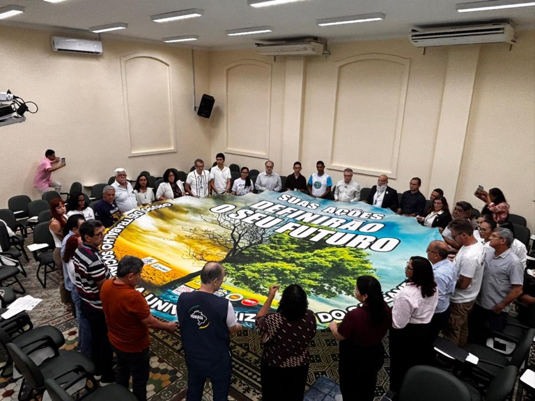Grupo de pessoas em círculo segurando uma grande faixa com mensagem ambiental no centro de uma sala de reuniões