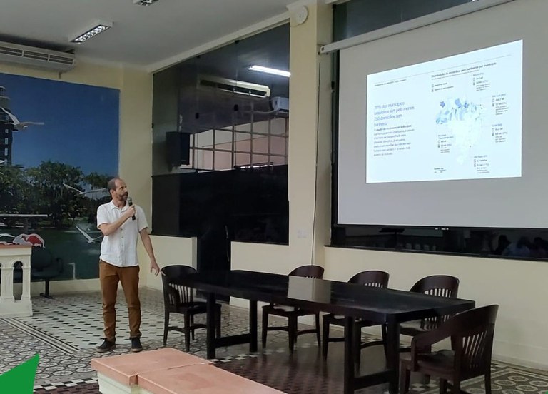 Apresentador com microfone mostra slide projetado em tela, em sala com mesa e cadeiras vazias.