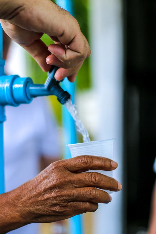 Mão abre torneira azul enquanto outra segura copo plástico sendo cheio com água potável