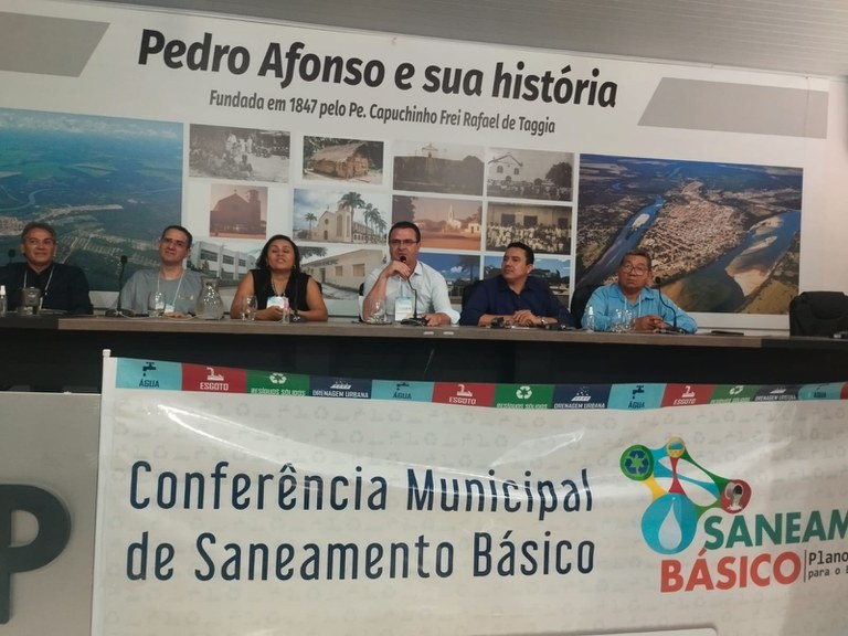 Mesa com sete pessoas em conferência de saneamento em Pedro Afonso; banner à frente e painel histórico da cidade ao fundo