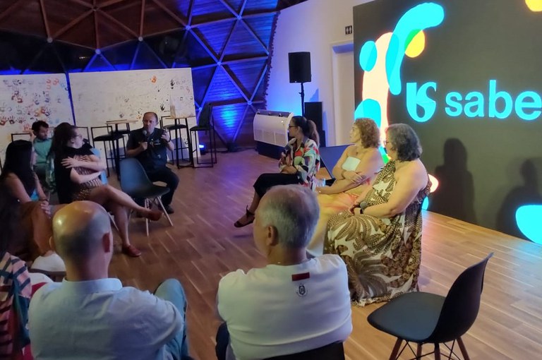 Roda de conversa em sala iluminada, com pessoas sentadas enquanto um participante fala e a tela exibe o logo “Saber”.