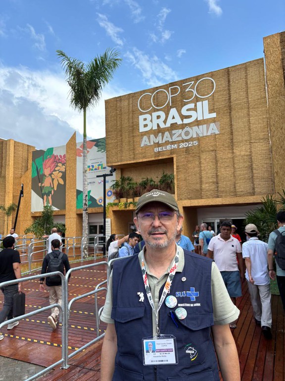 Presidente da Funasa com crachá e colete do SUS posa em frente ao pavilhão da COP30 em Belém, com pessoas circulando ao fundo.