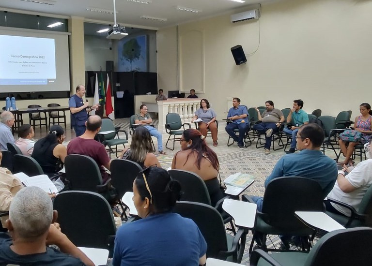 Grupo de pessoas reunidas em sala ampla, sentadas em círculo, ouvindo um palestrante que apresenta dados do Censo 2022 projetados na tela.
