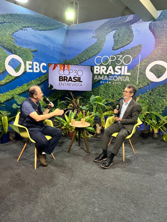 Duas pessoas sentadas em poltronas, conversando com microfones em cenário da COP30 com plantas ao redor.