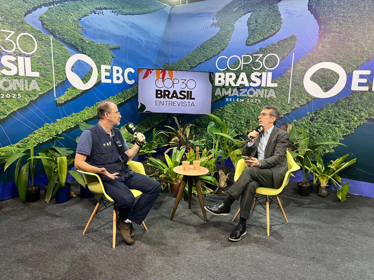 Entrevistador e entrevistado falam ao microfone em cenário da COP30, cercados por plantas e painel da Amazônia.