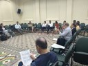 Reunião em roda com participantes sentados lendo documentos, em sala ampla com piso estampado.