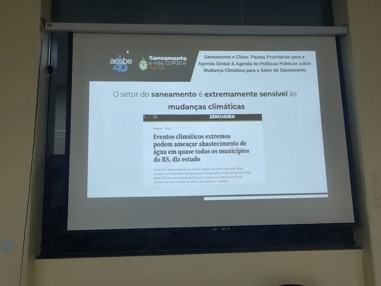 Tela de apresentação projeta notícia sobre impacto das mudanças climáticas no saneamento.