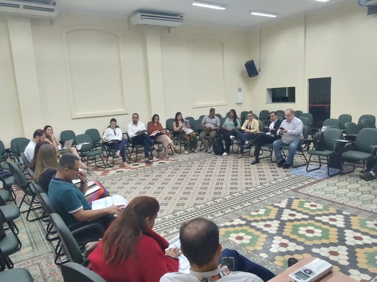 Grupo de participantes sentados em semicírculo discutindo documentos em sala ampla.